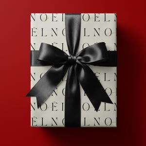 May include: A rectangular gift box wrapped in off-white paper featuring a repeating black serif typography pattern of the word NOËL. A wide, smooth black satin ribbon is tied in a large, centered bow around the exterior of the package.