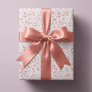 May include: A rectangular gift box wrapped in white paper featuring a scattered pattern of small pink, red, and orange hearts. A wide, smooth, solid rose gold satin ribbon is tied around the box in a cross pattern with a large bow at the center.