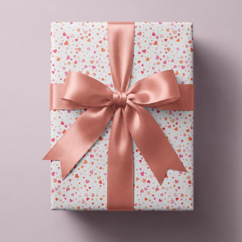 May include: A rectangular gift box wrapped in white paper featuring a scattered pattern of small pink, red, and orange hearts. A wide, smooth, solid rose gold satin ribbon is tied around the box in a cross pattern with a large bow at the center.