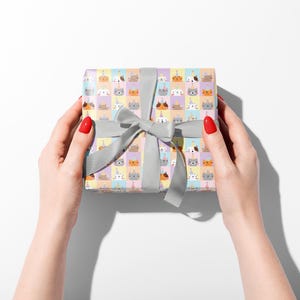 May include: A gift box wrapped in patterned paper with cartoon animal faces, including cats and unicorns, is held by two hands. The box is tied with a grey ribbon and bow. The background is white.