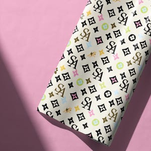 Könnte beinhalten: Cremefarbenes Geschenkpapier mit einem sich wiederholenden Muster aus schwarzen, braunen und goldenen Buchstaben und Symbolen. Der Hintergrund ist ein zartes Rosa. Das Papier ist gefaltet und zeigt das Muster auf der Vorderseite.