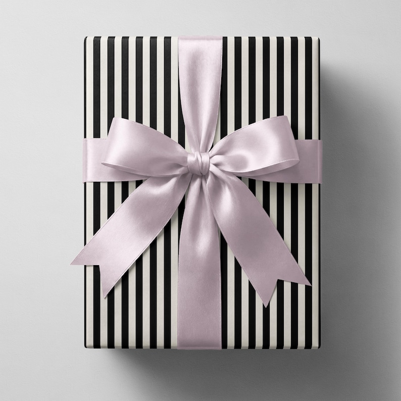 May include: A rectangular gift box wrapped in paper featuring a pattern of vertical black and white stripes. The box is tied with a wide, smooth, light lavender satin ribbon that is tied into a large, centred bow with two long tails.