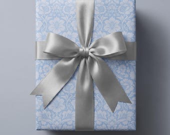 May include: A rectangular gift box wrapped in light blue paper featuring a white damask floral pattern. The package is tied with a wide, smooth, silver-colored satin ribbon that is arranged into a large, centered bow with two long tails.