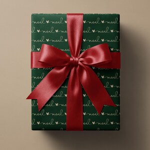 May include: A rectangular gift box wrapped in dark green paper featuring a repeating pattern of the word noel in cursive script interspersed with small hearts in a light beige color, tied with a wide, shiny, deep red satin ribbon bow.