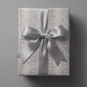 May include: A rectangular gift box wrapped in light gray and white paper featuring a repeating snakeskin pattern. The package is tied with a wide, smooth, solid silver satin ribbon that forms a large, centered bow with two long, pointed tails.
