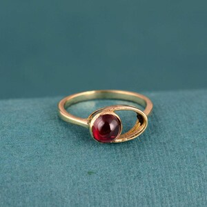 May include: A gold ring with a round, deep red gemstone. The ring has a unique design, with an open, oval-shaped setting around the gemstone. The band is a simple, polished gold. The ring is set against a teal background.