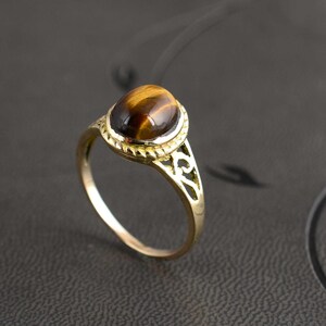 Puede incluir: Un anillo dorado con una piedra preciosa de ojo de tigre ovalada. El anillo tiene un diseño calado ornamental en la banda y un engaste decorativo. La piedra ojo de tigre muestra bandas marrones y doradas.