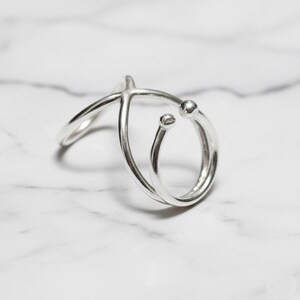Könnte beinhalten: Ein silberfarbener Ring mit einem einzigartigen, offenen Design. Der Ring hat ein gebogenes Band, das den Finger umschließt, mit zwei kleinen, kugelförmigen Akzenten. Das Design des Rings ist modern und minimalistisch.