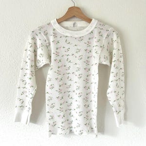 May include: A long-sleeve thermal shirt in white featuring a repeating pattern of small pink flowers and green leaves. The shirt has a ribbed texture, a crew neckline, and plain cuffs, hanging on a wooden hanger against a plain white wall.