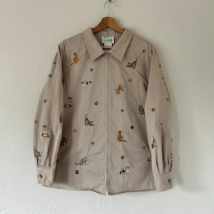 May include: A beige long-sleeve zip-up jacket featuring an all-over embroidered pattern of various cats, yarn balls, and paw prints. The jacket has a classic collar, buttoned cuffs, and a central zip closure, showcasing a light tan fabric base.