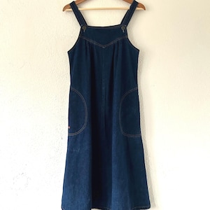 Può includere: Un abito scamiciato in denim blu scuro con scollo quadrato, spalline larghe con chiusura a bottoni e due ampie tasche laterali. Il capo presenta una cucitura a forma di V sul petto ed è appeso a una gruccia di legno.