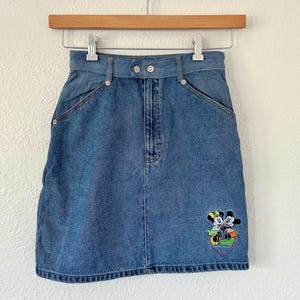 May include: A blue denim skirt with a classic design. Features a colorful embroidered image of Mickey and Minnie Mouse on the front. The skirt has a button and zipper closure, front pockets, and decorative stitching. A vintage clothing item.