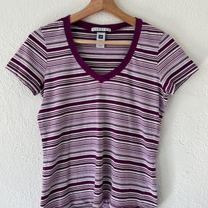 以下が含まれることがあります： 紫と白のストライプ柄の半袖VネックTシャツ。木製のハンガーにかかっています。ブランド名「STRETCH」がネックラインに表示されています。