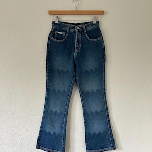 May include: High-waisted blue denim jeans featuring a repeating horizontal zigzag pattern in a slightly darker shade of blue. The jeans have a classic five-pocket design, a button and zipper fly closure, and a straight-leg cut that flares slightly.