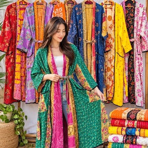 May include: A long, open-front robe featuring a patchwork design with vibrant green, pink, and gold floral patterns. The garment includes long sleeves, a matching tie belt, and black trim along the edges and pockets, displayed among other similar robes.