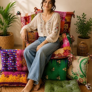 Puede incluir: Una blusa beige de manga larga con bordados florales y escote con lazos, combinada con vaqueros azul claro. La blusa se lleva puesta mientras se está sentado sobre un cojín de patchwork colorido, rodeado de otros cojines de suelo cuadrados.