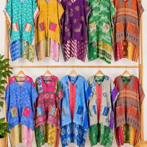 May include: Ten sleeveless tunics with V-necklines and two front pockets, displayed on a wooden rack. Each garment features a unique combination of colorful, patterned fabrics with visible stitching, varying in length with a high-low hemline.