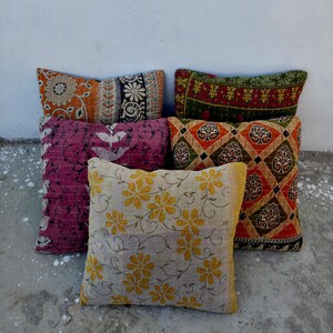 May include: Five square throw cushions featuring various patterns and colours. One is cream with yellow flowers, one is magenta with white leaf motifs, one is orange with black floral designs, one is green with red patterns, and one has a diamond print.