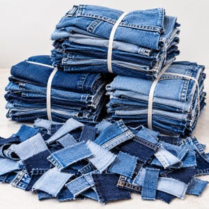 Könnte beinhalten: Drei Stapel gefalteter blauer Denim-Stoffreste, die mit weißen Bändern zusammengehalten werden, sowie ein loser Haufen kleinerer rechteckiger Denim-Stücke in verschiedenen Blautönen, von hell bis dunkel, mit deutlich sichtbaren ausgefransten Kanten.