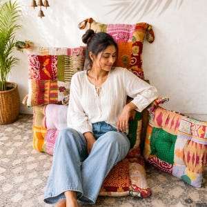 May include: A white long-sleeved blouse with a tie-front neckline and vertical pleats, paired with light blue denim jeans. The clothing is positioned against a collection of square floor cushions featuring a colorful patchwork pattern.