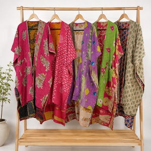 Puede incluir: Cinco kimonos abiertos colgados en un perchero de madera. Las prendas presentan varios patrones coloridos, incluyendo estampados florales y geométricos en tonos rosa, verde, morado, amarillo y beige, con costuras visibles de estilo kantha.