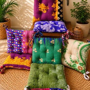 May include: A collection of square floor cushions featuring various colorful patterns, including floral, polka dot, and botanical prints. Each cushion is tufted with visible stitching and includes fabric ties at the corners for securing in place.