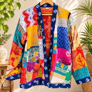 May include: A long-sleeved jacket featuring a vibrant patchwork design with various colourful fabric squares in shades of red, yellow, blue, and pink. The garment includes a dark blue trim with a floral pattern, two front pockets, and visible stitching.