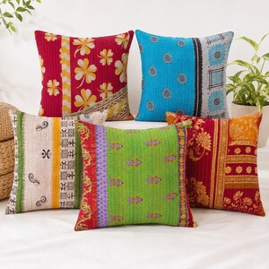 Könnte beinhalten: Eine Sammlung von fünf quadratischen Dekokissen mit verschiedenen bunten Mustern. Die Kissen zeigen leuchtende rote, blaue, grüne und cremefarbene Stoffe mit floralen, geometrischen und Paisley-Drucken, alle mit sichtbaren Steppnähten im Kantha-Stil.