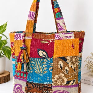 May include: A quilted tote bag featuring a patchwork design of various floral and abstract patterns in vibrant colours including red, blue, yellow, and purple. The bag has two matching fabric handles and a decorative multi-coloured fabric tassel.