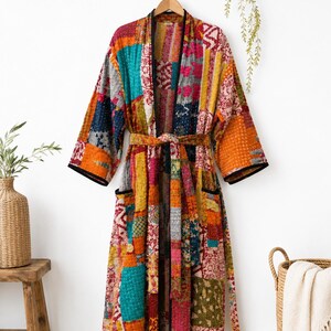 May include: A long-length robe featuring a vibrant patchwork design with various colorful patterns in shades of orange, red, yellow, blue, and pink. The garment includes a matching tie belt, two front pockets, and black trim along the edges.