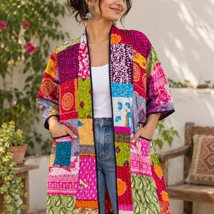 Könnte beinhalten: Eine langärmelige, vorne offene Jacke mit einem lebendigen Patchwork-Design aus verschiedenen bunten Mustern in Rosa, Grün, Gelb und Blau. Das Kleidungsstück verfügt über zwei Vordertaschen und ist an den Kanten mit einem schwarzen Saum versehen.