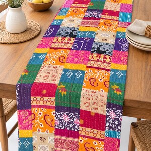 Puede incluir: Un camino de mesa rectangular con un diseño de retazos de varios cuadrados de tela en colores vibrantes que incluyen amarillo, magenta, azul cerceta y morado. La tela muestra diversos patrones florales, de cachemira y geométricos con costuras blancas visibles.