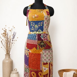 Könnte beinhalten: Eine Patchwork-Schürze aus verschiedenen Stoffquadraten mit Blumen-, geometrischen und Paisley-Mustern in Gelb-, Blau-, Rot- und Violetttönen. Die Schürze hat ein Nackenband und ist mit sichtbaren weißen Steppnähten versehen.