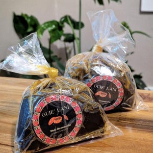 May include: Two clear plastic bags, each containing a dark brown, rectangular food item. The bags are tied with yellow twist ties. Each bag has a circular label with the text "GUIE' LANI" and a red floral border.