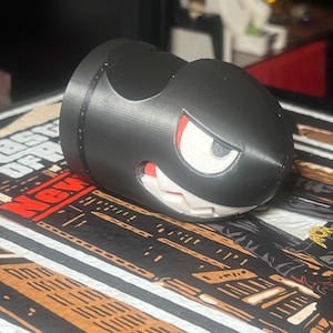 Puede incluir: Una figura negra de Bala de Cañón con boca blanca, ojos rojos y un ojo blanco. La figura es un artículo impreso en 3D. La figura descansa sobre una superficie estampada con colores rojo, negro y marrón.