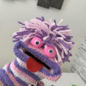 May include: A handmade sock puppet with a whimsical design. The puppet features a striped body in shades of pink, white, and purple. It has large googly eyes, a red felt tongue, and a fluffy yarn hair in purple and pink.