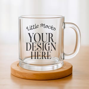 May include: A clear glass mug with a handle, on a round wooden coaster. The mug has the text "Little Mocks YOUR DESIGN HERE" in black. The mug is empty and sits on a light brown wooden surface. The coaster is made of wood.