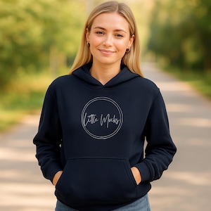May include: A navy blue hooded sweatshirt with a front pocket and a drawstring hood. The text "Little Mocks" is displayed in a white, circular design on the chest. The person is wearing the sweatshirt outdoors.