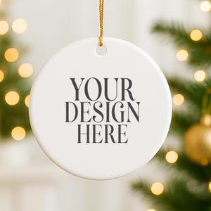 Może przedstawiać: Biała, okrągła ozdoba choinkowa z czarnym napisem "YOUR DESIGN HERE". Ozdoba zawieszona jest na złotym sznurku. Tło przedstawia rozmyte drzewko bożonarodzeniowe i światełka.