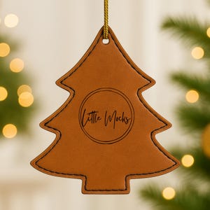 Puede incluir: Adorno navideño de cuero marrón con costuras negras y una cuerda dorada para colgar. El adorno tiene las palabras "Little Mocks" en un círculo en el centro. El fondo está borroso con luces navideñas.