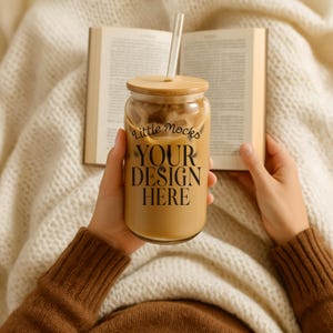 Puede incluir: Un vaso de vidrio lleno de una bebida de café, con tapa de madera y pajita de vidrio. El vaso tiene el texto "Little Mocks YOUR DESIGN HERE". Se sostiene frente a un libro abierto y una manta blanca.