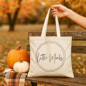 以下が含まれることがあります： 「Little Mocks」の文字が円形デザインでプリントされたクリーム色のトートバッグ。バッグは手で持たれています。オレンジ色のカボチャと白いカボチャが、チェック柄のブランケットが敷かれた木製のベンチに置かれています。秋のシーン。