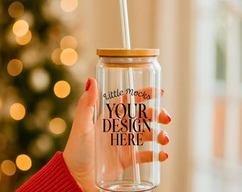 Puede incluir: Un vaso de vidrio transparente con tapa de bambú y pajita blanca. El vaso tiene el texto "Little Mocks YOUR DESIGN HERE" impreso en la parte delantera. La persona que sostiene el vaso lleva un jersey rojo.