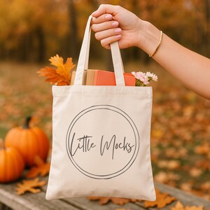 以下が含まれることがあります： クリーム色のトートバッグ。 黒の手書き風フォントで「Little Mocks」の文字が二重の円で囲まれている。 バッグには本、花、紅葉が入っている。 背景にはカボチャが見える。