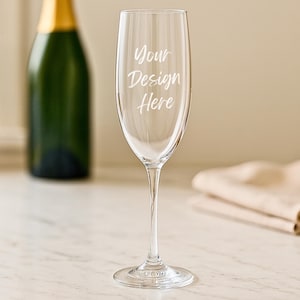 May include: A clear champagne flute with the text "Your Design Here" etched on the glass. The flute has a long stem and a wide bowl. A green champagne bottle is visible in the background.