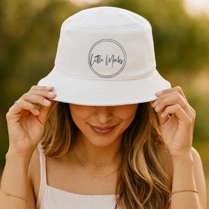 May include: White bucket hat with a circular logo that reads "Little Mocks" in a handwritten font. The hat is being held by a person. The background is blurred, with a natural, outdoor setting.