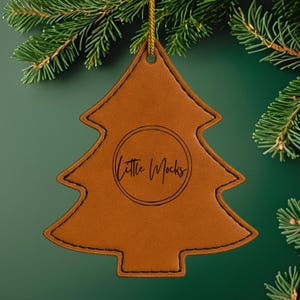 Puede incluir: Adorno navideño de cuero marrón con costuras negras y cordón dorado para colgar. El adorno tiene el texto "Little Mocks" en un círculo. Cuelga sobre un fondo verde con agujas de pino.