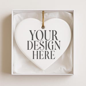 May include: A white heart-shaped ornament with the text "YOUR DESIGN HERE" in dark gray. The ornament hangs from a gold string and is presented in a white box with tissue paper.