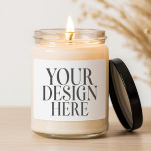 Peut inclure: Une bougie allumée dans un pot en verre transparent avec un couvercle noir. La bougie a une étiquette blanche avec le texte "YOUR DESIGN HERE" en noir. La bougie est sur une surface en bois.