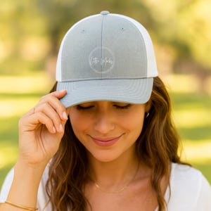 May include: A light gray and white trucker hat with the text "Little Mucks" in a white circle. The hat has a curved brim and a mesh back. The person in the image is wearing the hat.